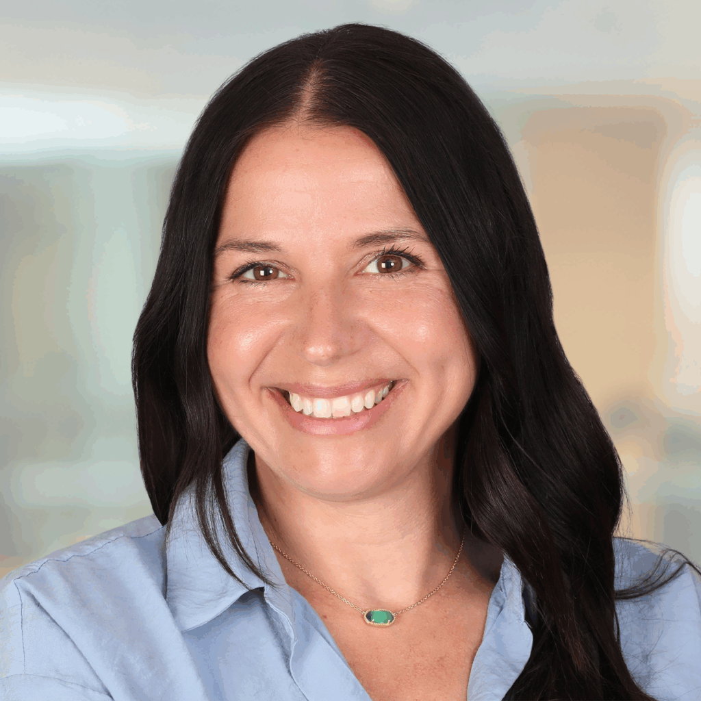 Professional headshot of Christine Molbury, a woman with long dark hair, wearing a light blue blouse and a green stone necklace, smiling warmly in front of a softly blurred office background.,