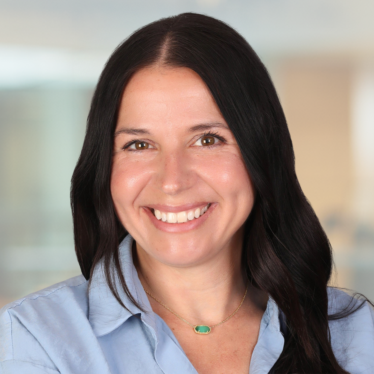 Professional headshot of Christine Molbury, a woman with long dark hair, wearing a light blue blouse and a green stone necklace, smiling warmly in front of a softly blurred office background.,